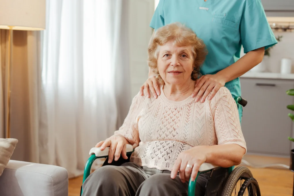 Elderly woman in wheelchair with caregiver’s supportive hands on shoulders.