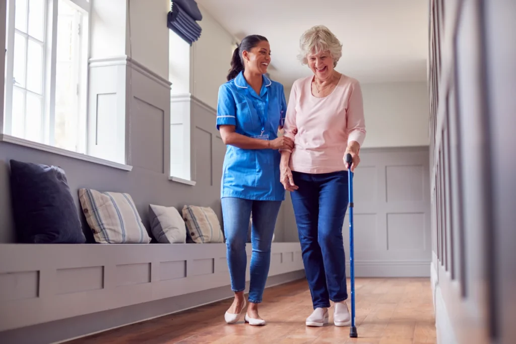 Caregiver assisting elderly woman walking hallway.