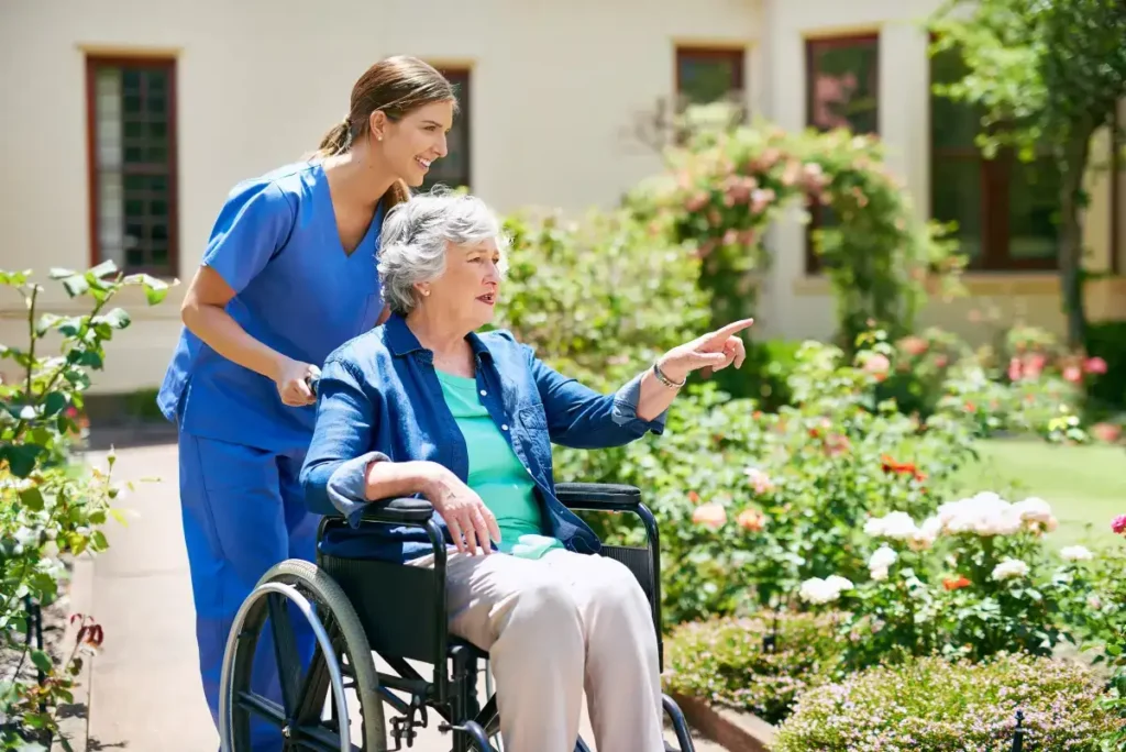Caregiver pushing elderly woman in wheelchair through garden.