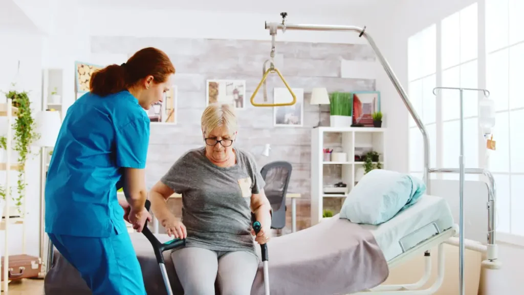 Caregiver assisting elderly woman standing bedside.